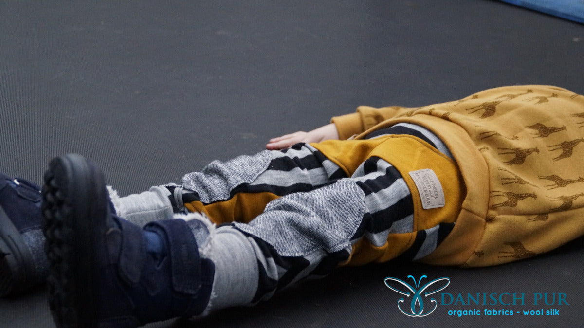 Kind auf Trampolin liegend in Giraffe Pullover aus Wollfrottee und gestreifter Wolljersey Leggins.