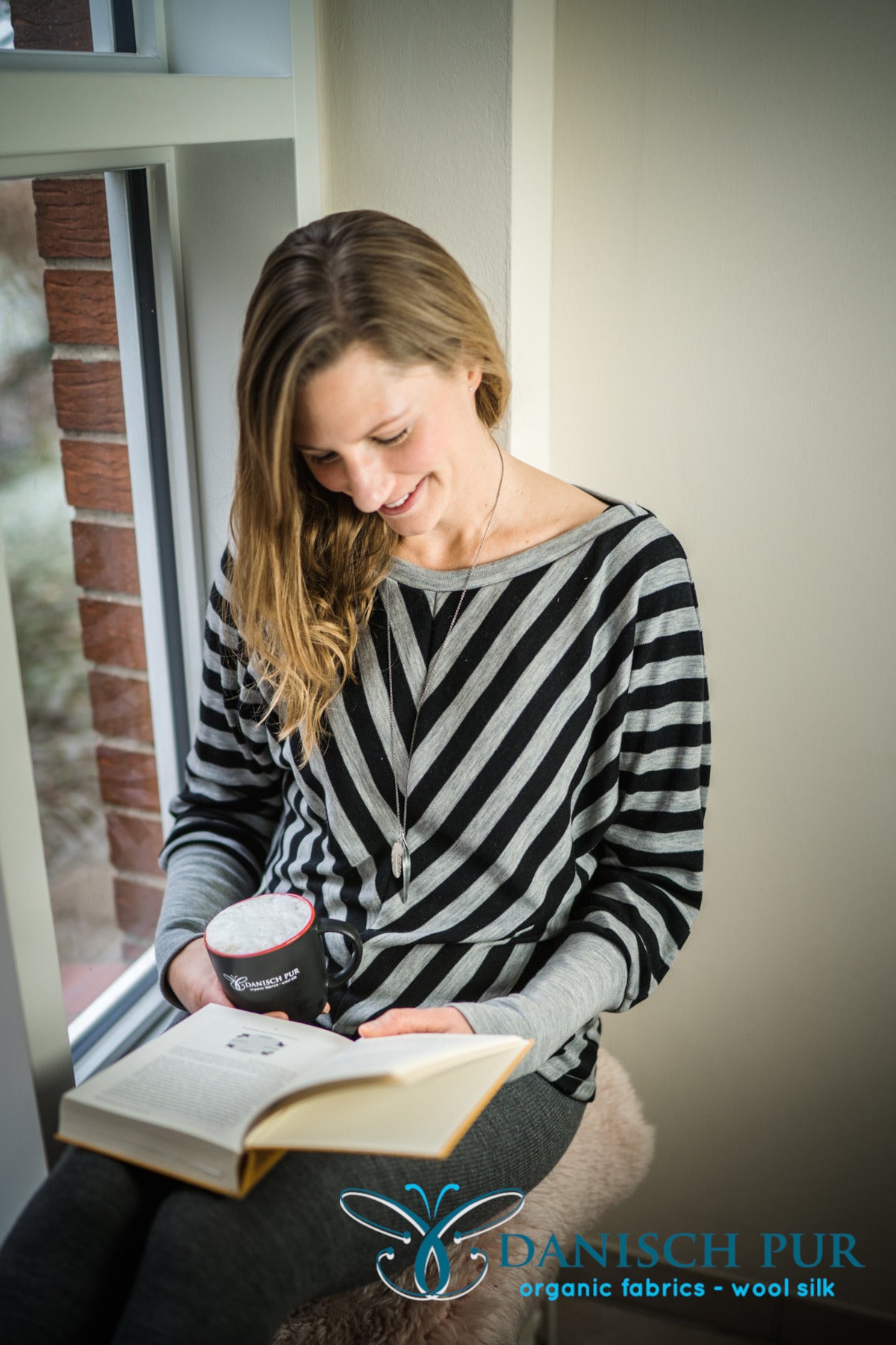 Am Fenster sitzende Frau in gestreiftem Wolljersey Pullover mit Buch und Kaffeetasse in der Hand.