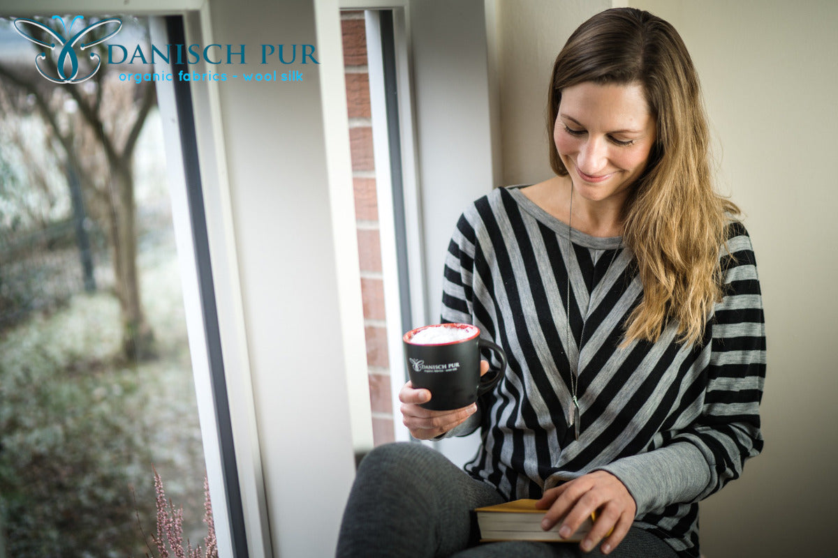 Frau auf Fensterbank sitzend in gestreitem Wolljersey Pullover. In den Händen hält sie einen Kaffee und ein Buch.
