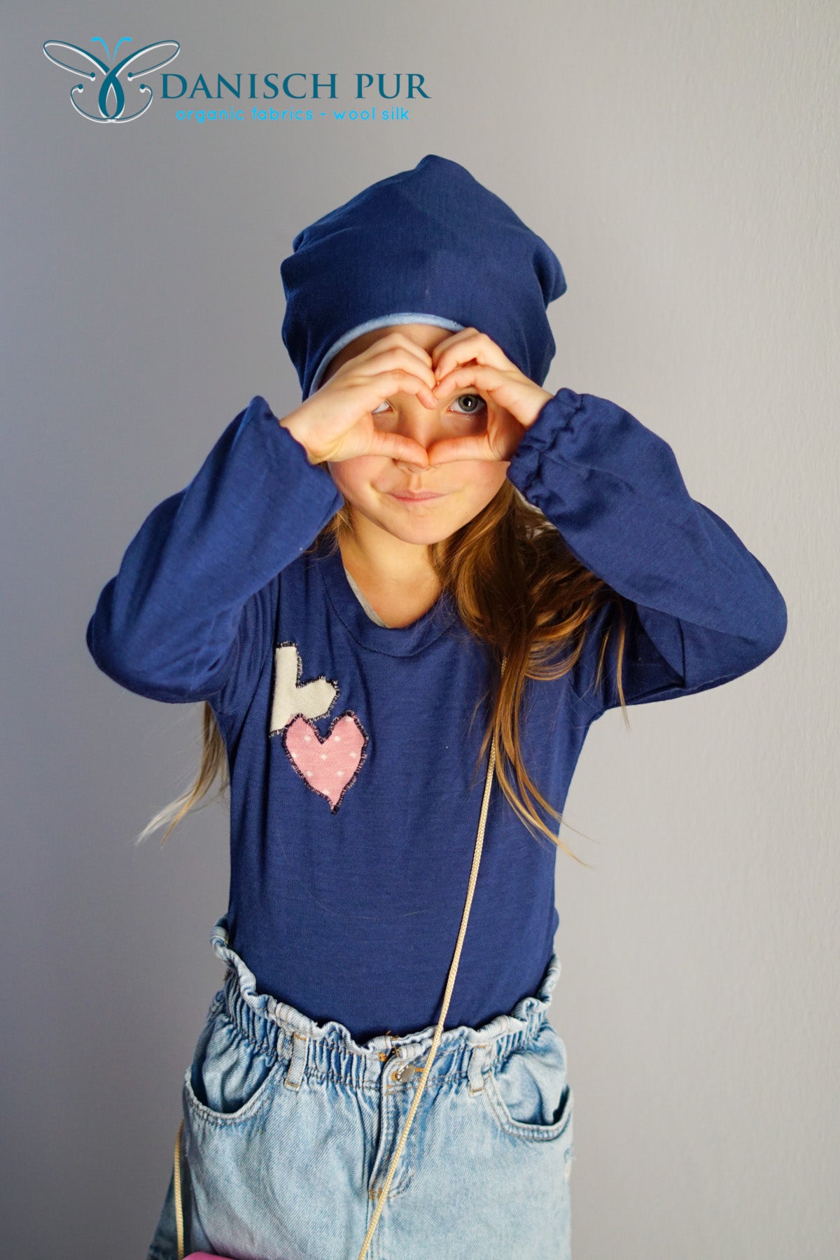 Mädchen in Shirt und Mütze aus Wolljersey faltet Hände zu einer Herzform zusammen vor dem Gesicht.